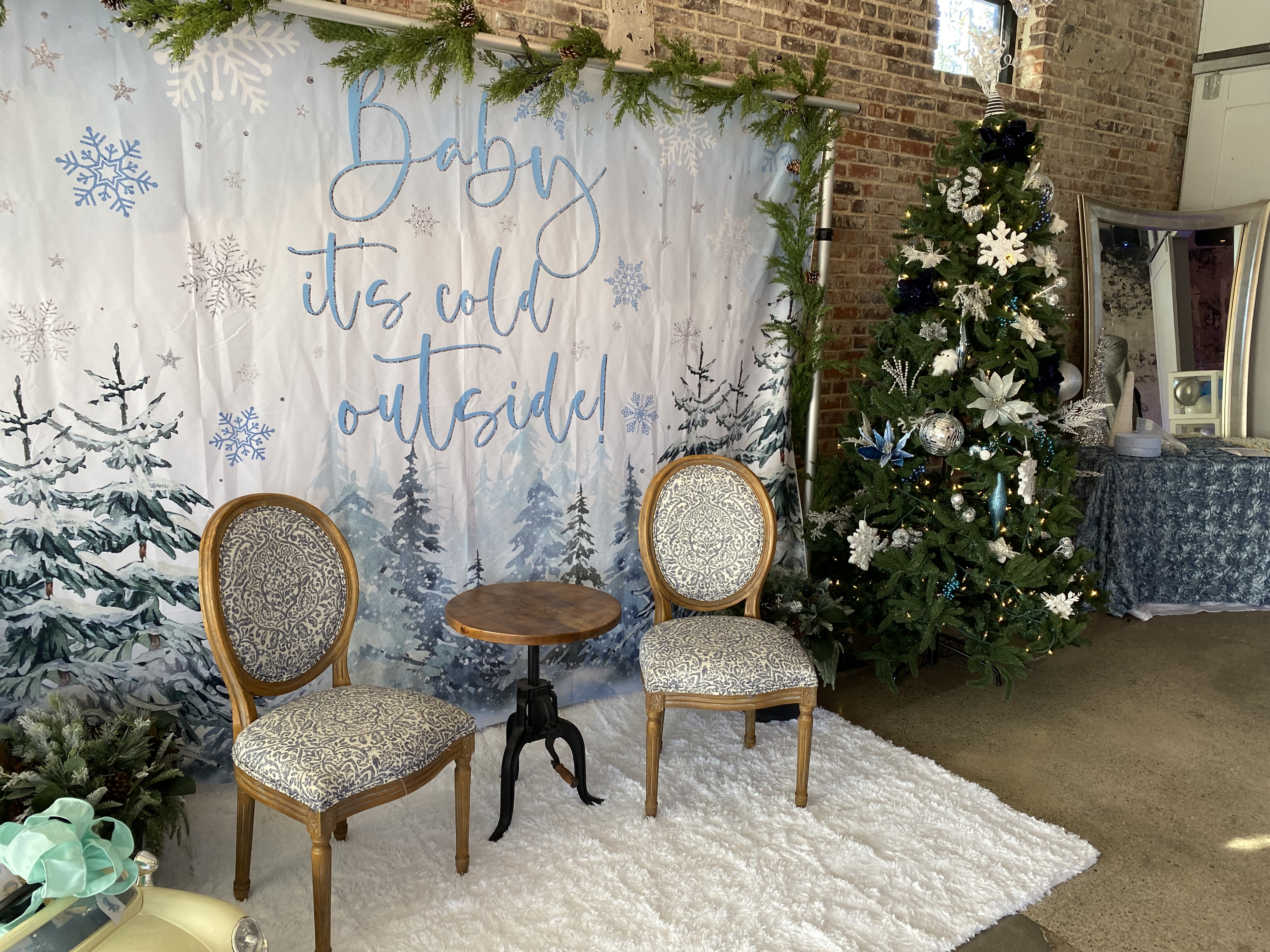 Winter-themed indoor setup with a backdrop reading 'Baby it's cold outside,' two vintage-style chairs, a small wooden table, and a decorated Christmas tree with white and blue ornaments.