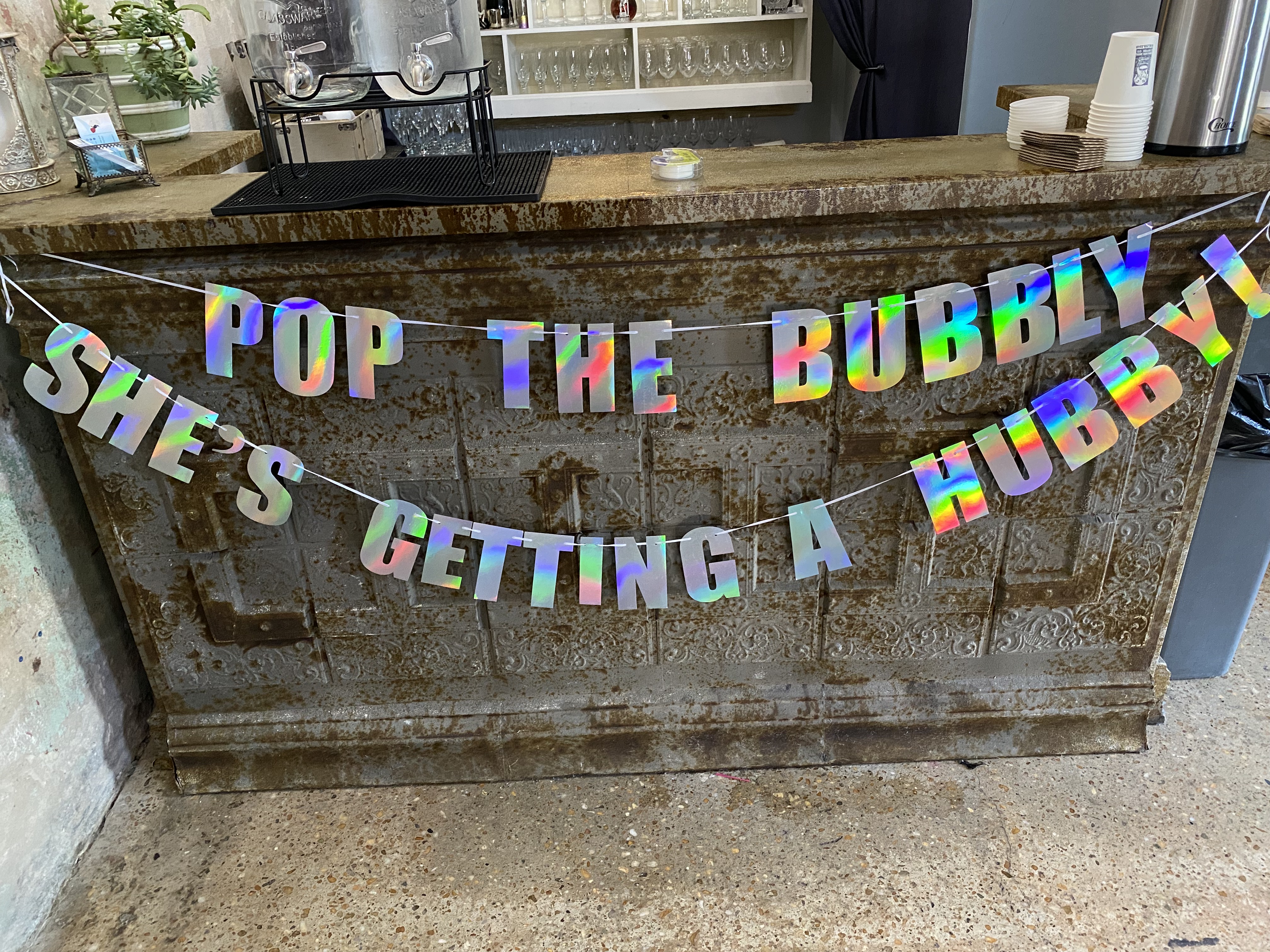 A rustic bar counter with a holographic banner that reads 'POP THE BUBBLY, SHE’S GETTING A HUBBY!' Glass dispensers, cups, and bar accessories are on the counter.