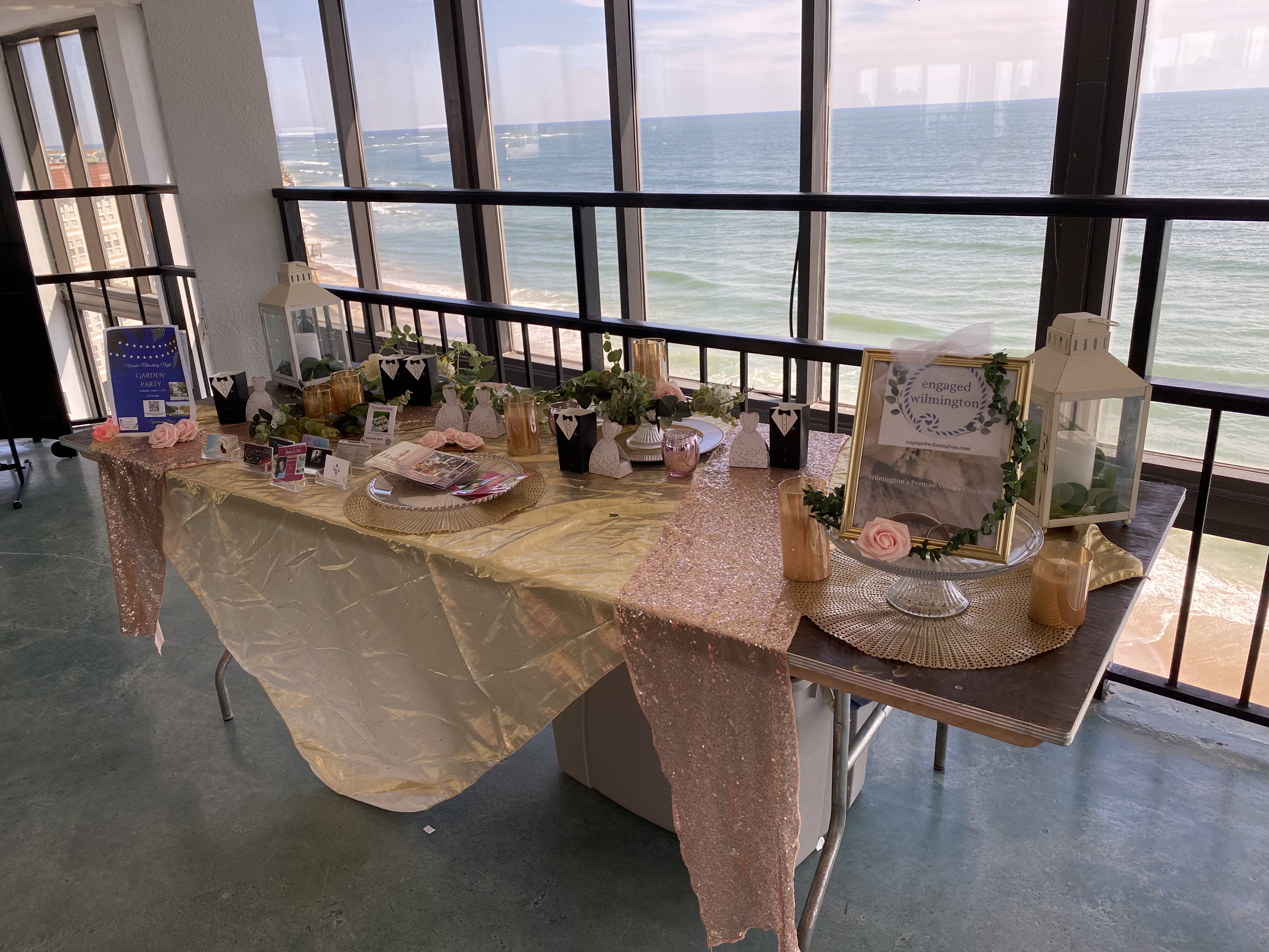 A bridal shower table decorated with gold and rose-gold tablecloths, lanterns, small tuxedo and dress gift boxes, and floral accents. The setup overlooks a scenic ocean view through large windows.